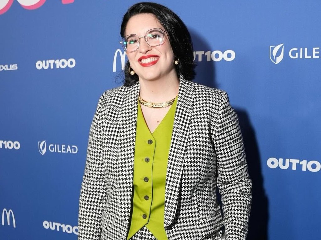 Kayla Gagnet smiling on the red carpet at Out Magazine's Out100 30th anniversary event, wearing a houndstooth suit and green vest.